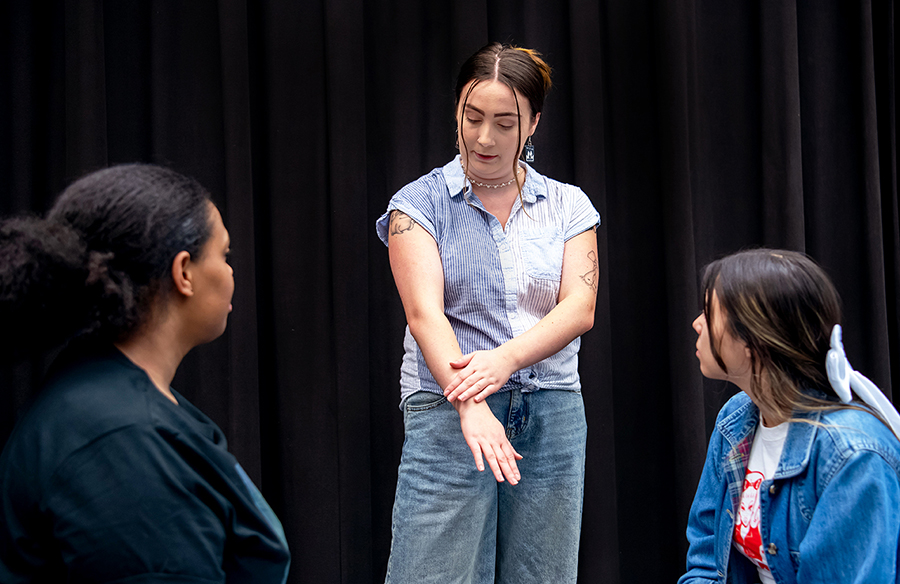 An intimacy certificate student leads a workshop with undergraduate theatre students. She is demonstrating touch through placing her hands on her forearms.