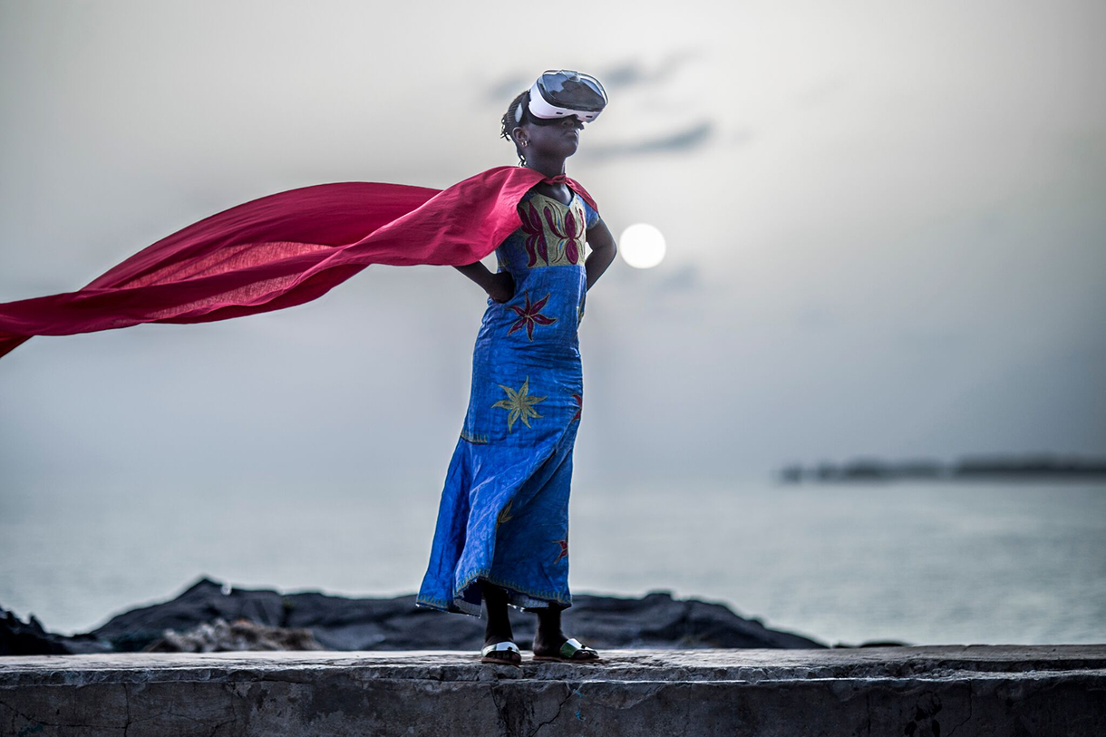 child wearing VR headset standing on beach with red cape blowing in wind
