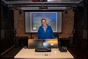 With a backdrop of a large screen, David Worrall stands behind computer screen and is surrounded by speakers .in Immersive lab