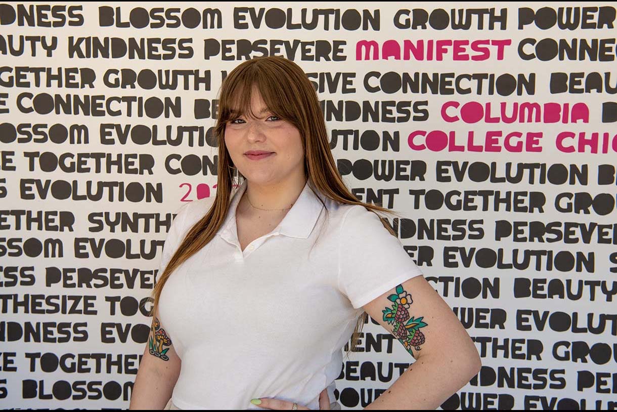 Miranda Prater posing in front of signage created for Columbia's annual Manifest celebration; sign includes unique font with words such as "Connection" "Kindness," and "Growth."