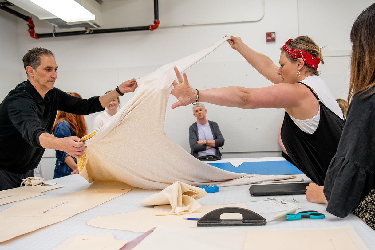 two people draping fabric at table with person observing in background