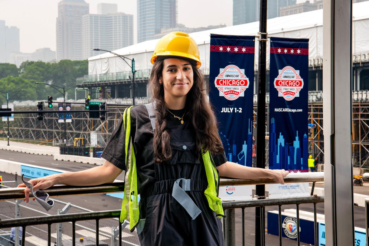 portrait of yasmine mifdal wearing hard hat with nascar race track being set up in background
